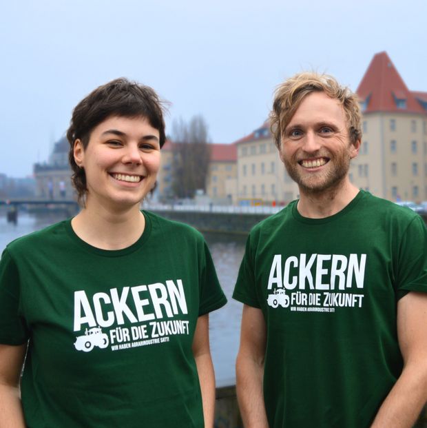 Flaschengrüne T-Shirts in geradem und tailliertem Schnitt mit weißem Text-und-Bild-Motiv: Ackern für die Zukunft – Wir haben Agrarindustrie satt. Neben dem Schriftzug ist klein ein moderner Traktor abgebildet.